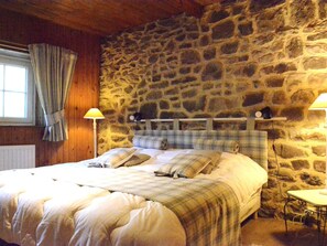 A bedroom with a stone wall, a bed with plaid bedding, two bedside lamps, and a window with curtains. - Holiday Home in Viroinval With Sauna (Viroinval)