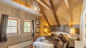 A bedroom with a wooden ceiling, a bed with a patterned bedspread, two bedside tables with lamps, a bench, and a radiator.