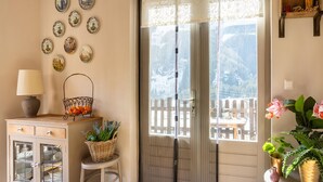 A room with a wooden cabinet with glass doors, a wicker basket on a stool, a window with a view of a wooden fence and mountains, and a table with plants.