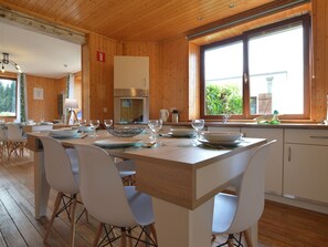 Una cocina moderna con paredes de madera, un ventanal grande y una zona de comedor con sillas blancas y una mesa de madera preparada para cuatro.