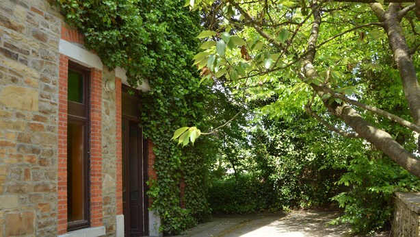 A stone building with a brick facade, ivy-covered walls, and a narrow pathway leading through a lush green area. - Family Villa in Ferrieres (Ferrieres)