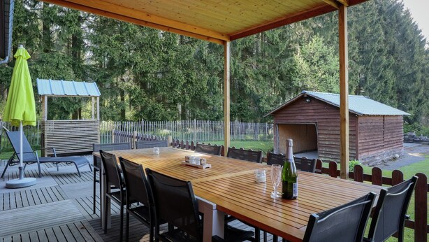 A covered outdoor seating area with a wooden table and chairs, a small gazebo, and a wooden cabin in a wooded setting.
