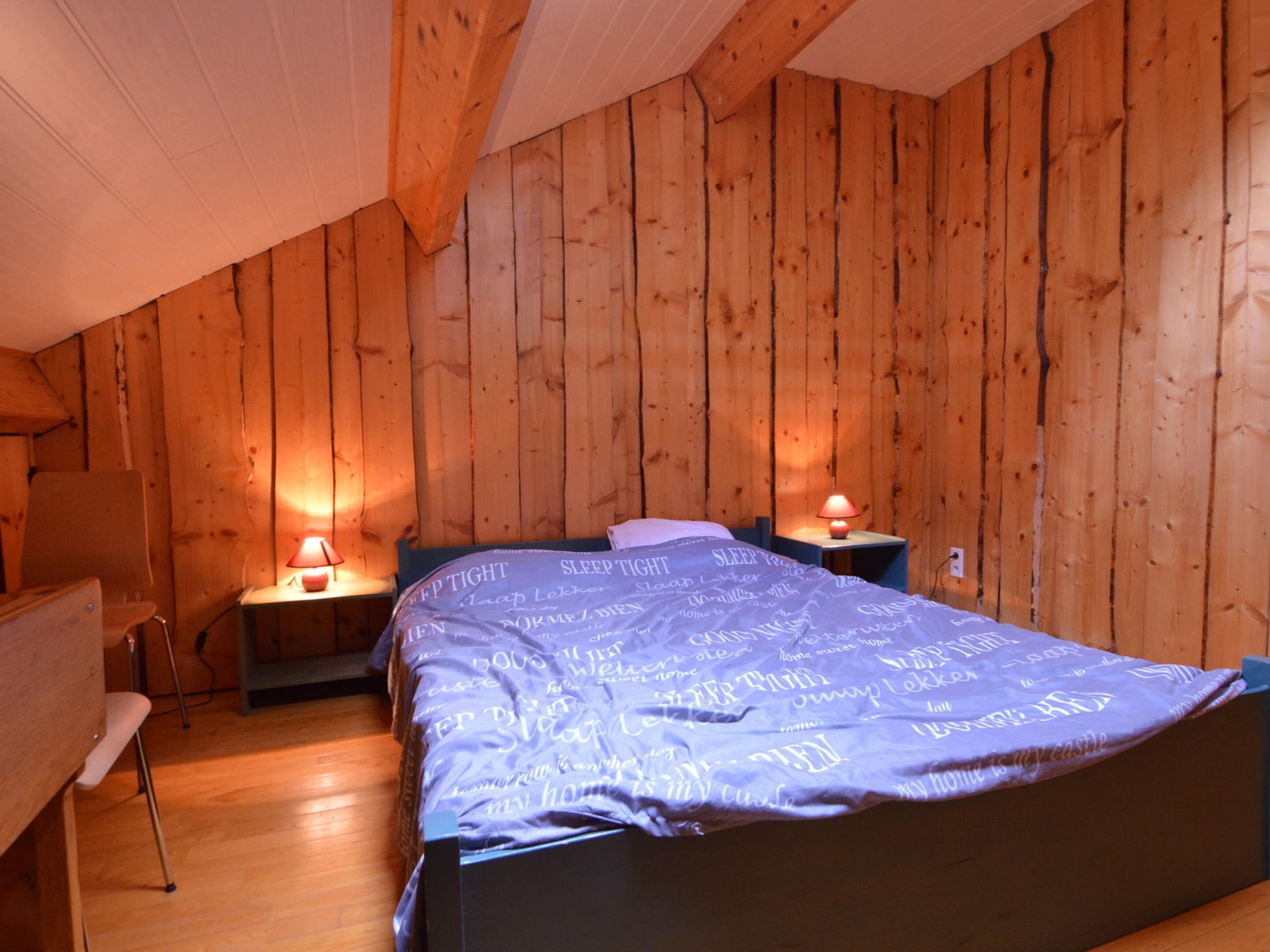 A bedroom with a wooden sloped ceiling, a bed with a blue blanket, two bedside tables with lamps, and a chair.