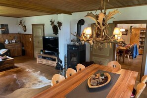 Ein rustikaler Essbereich mit einem Holztisch, Stühlen und einer dekorativen Mittelstück. Im Hintergrund sind ein Kamin und ein Fernseher zu sehen.