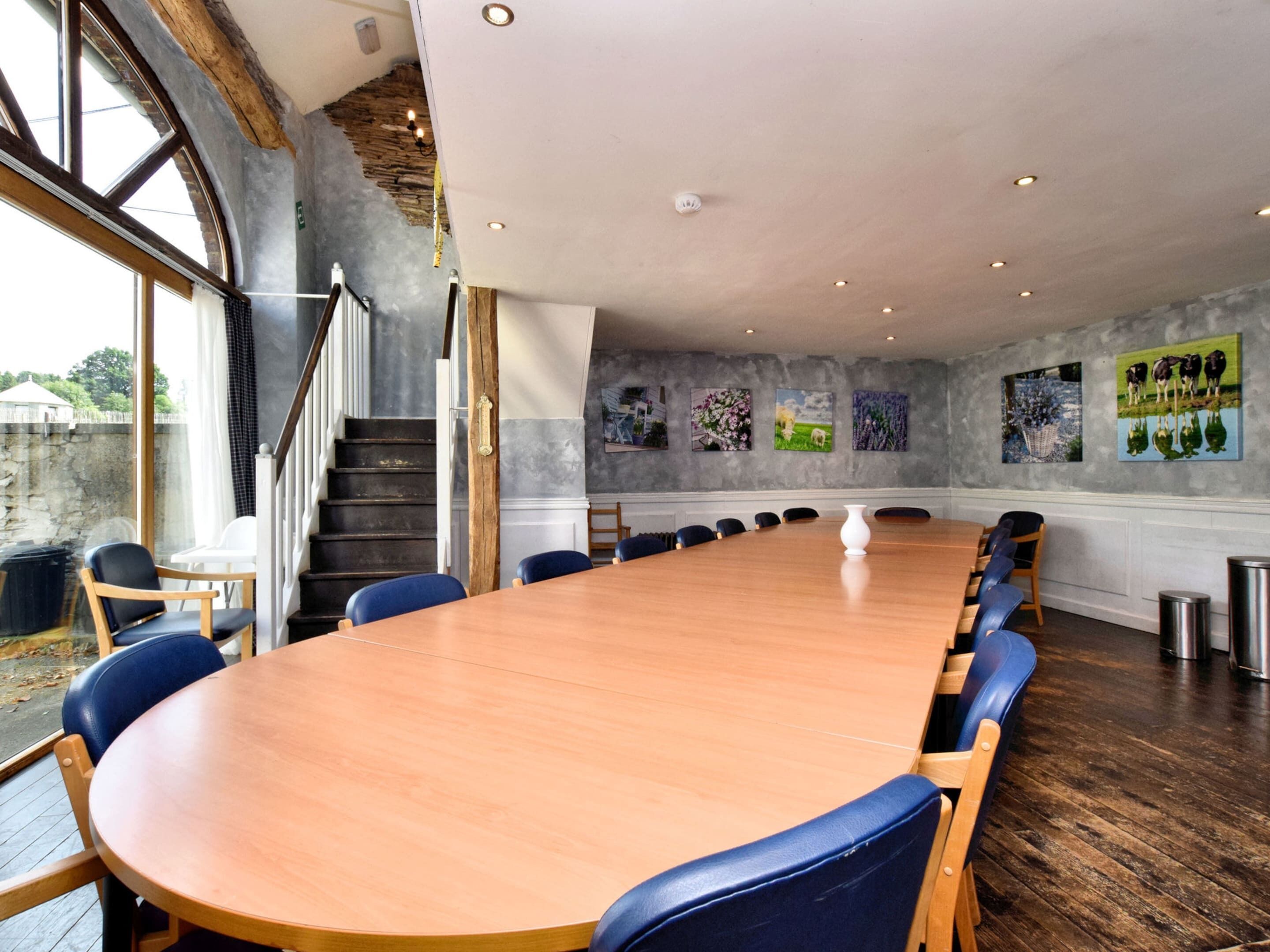 A conference room with a long wooden table, blue chairs, and a staircase leading to a balcony.