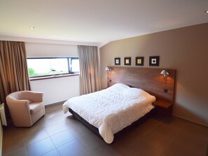 Een moderne slaapkamer met een groot bed, een stoel, een raam met gordijnen en een wand met ingelijste foto's.