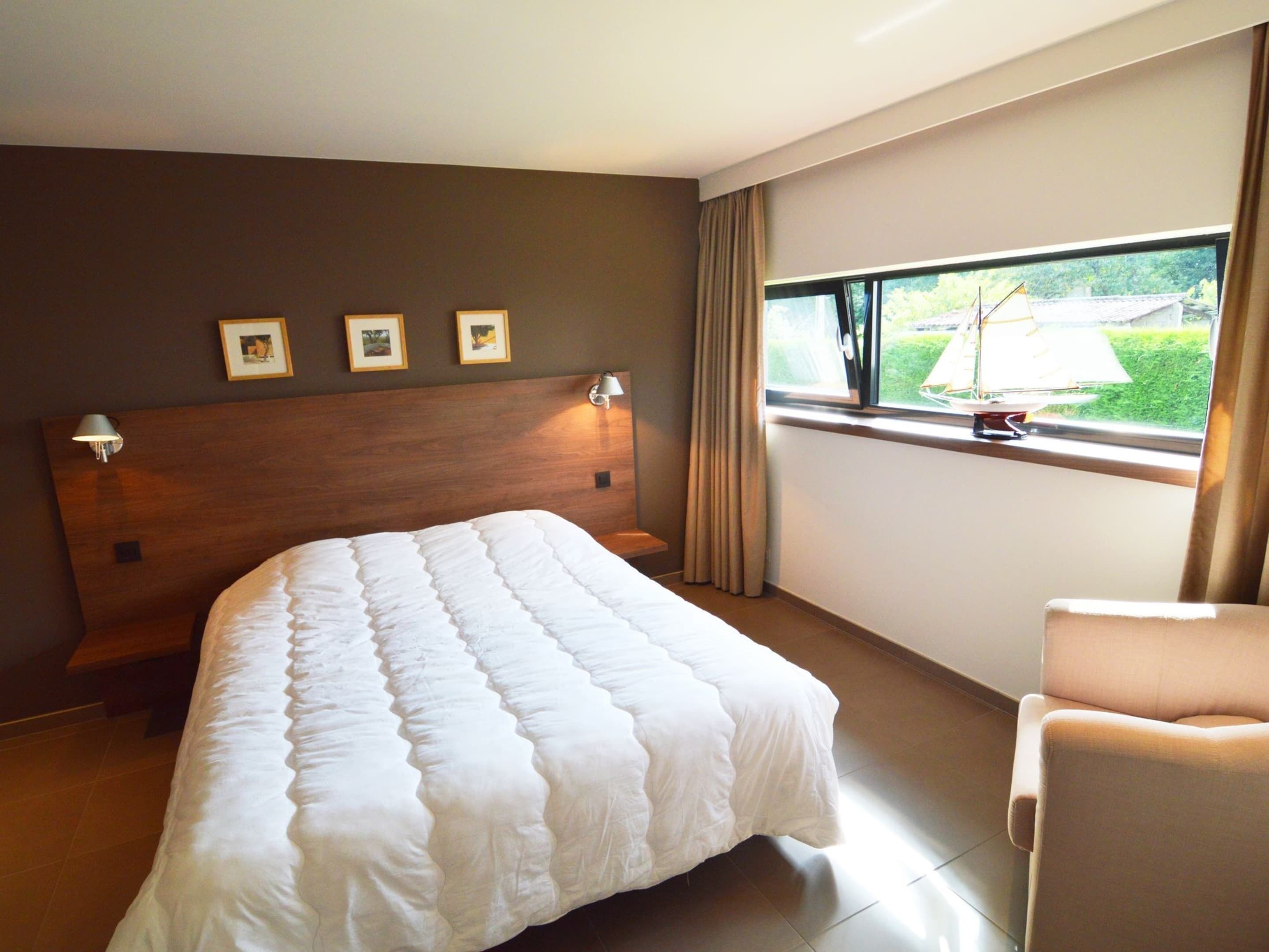 Un cuarto de hotel con una cama grande, una silla, un ventanal con vista a follaje, y tres cuadros en la pared.