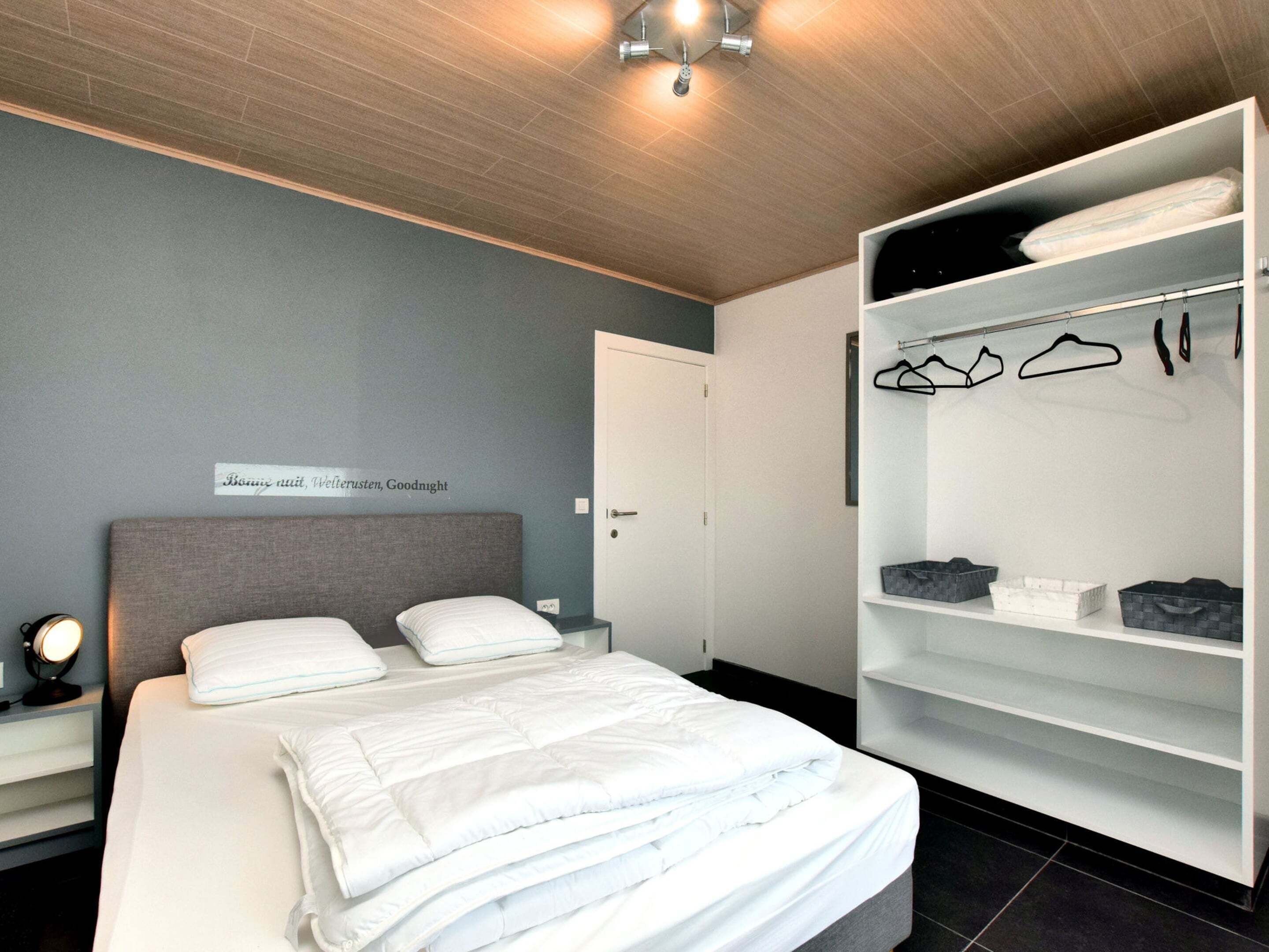 A modern bedroom with a neatly made bed, a bedside table with a lamp, a wardrobe with hanging clothes, and a sign on the wall.