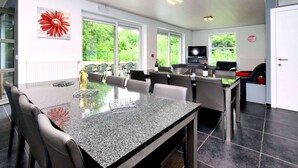 A modern kitchen with a granite countertop, dining area with chairs, and large windows offering a view of greenery. - Luxurious Home With Sauna (Somme-Leuze)