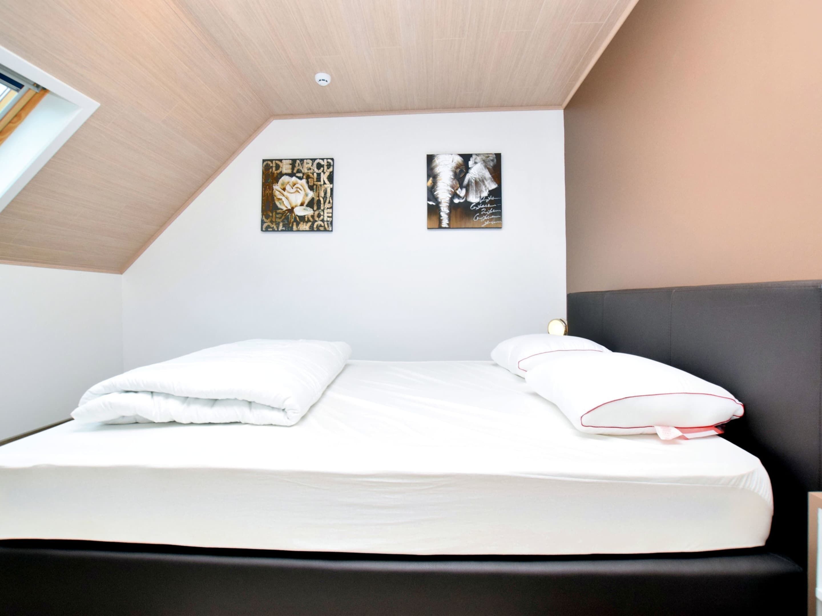 A neatly made bed with white linens in a modern room with a sloped ceiling and two wall-mounted artworks.
