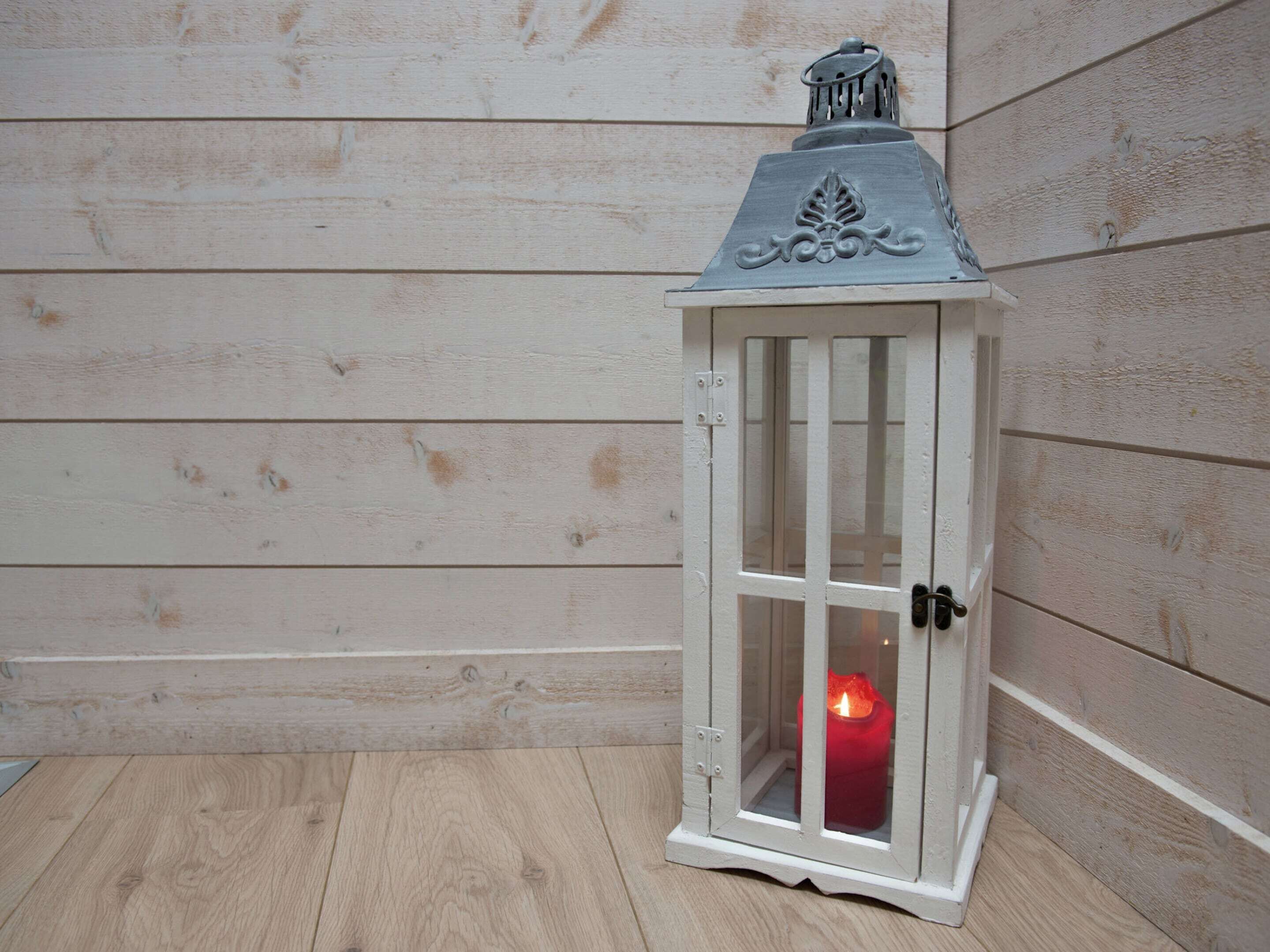 Une lanterne blanche contenant une bougie, posée sur un plancher en bois, contre un mur en bois.