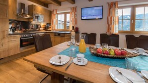 A modern kitchen with wooden cabinets, a built-in oven, and a microwave. There is a dining table set with plates, glasses, and a fruit basket. - Apartment in Kaprun With Ski-in/ski-out Access (Kaprun)