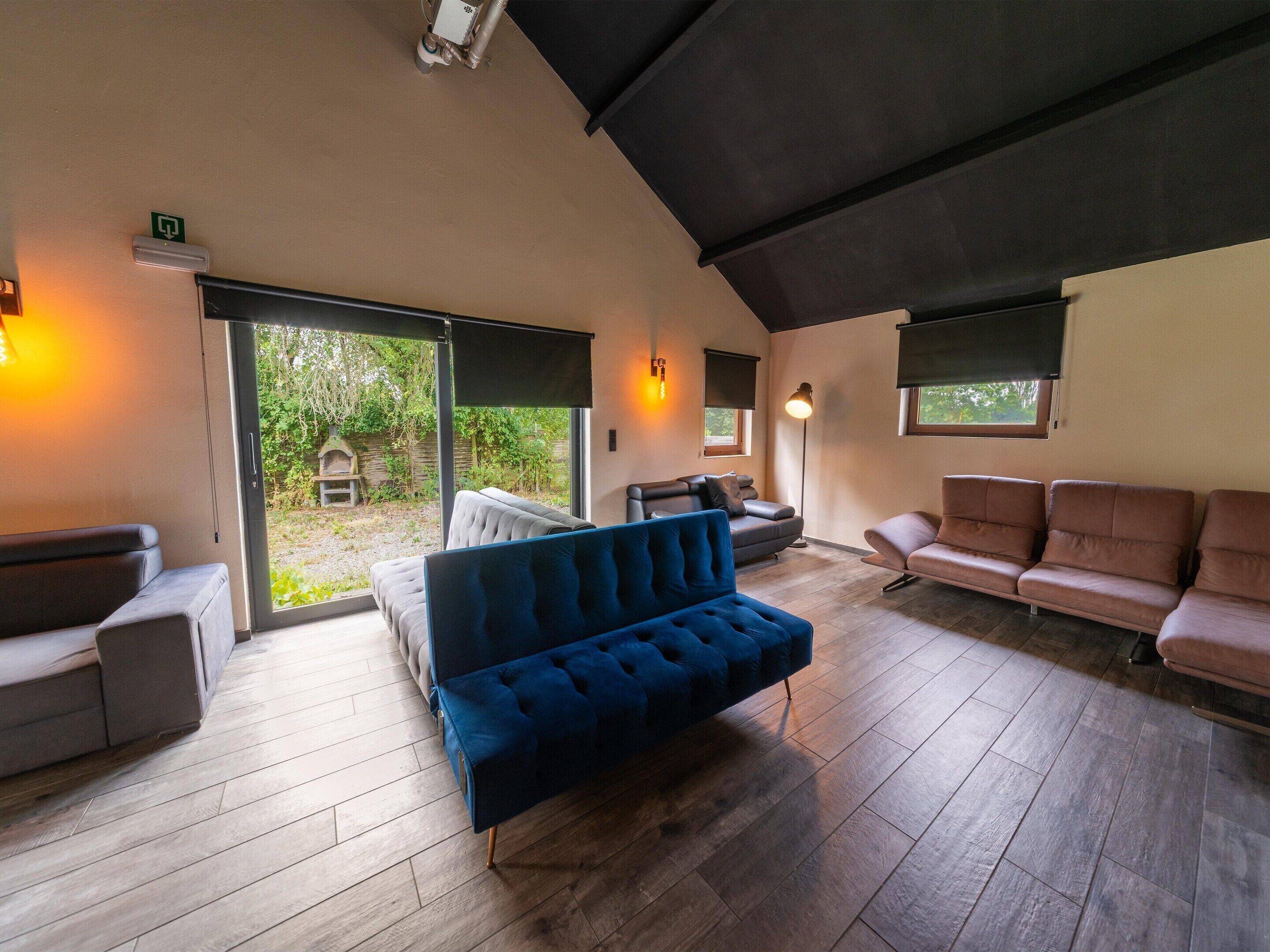 A spacious living area with a blue sofa, a grey sofa, and a brown sofa. There are windows with blinds, a fireplace, and a ceiling light fixture.