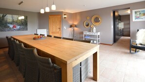 Un vaste espace dédié à la salle à manger, avec une longue table en bois, des chaises en osier, des luminaires suspendus et une vue sur une forêt, visible à travers une grande fenêtre.