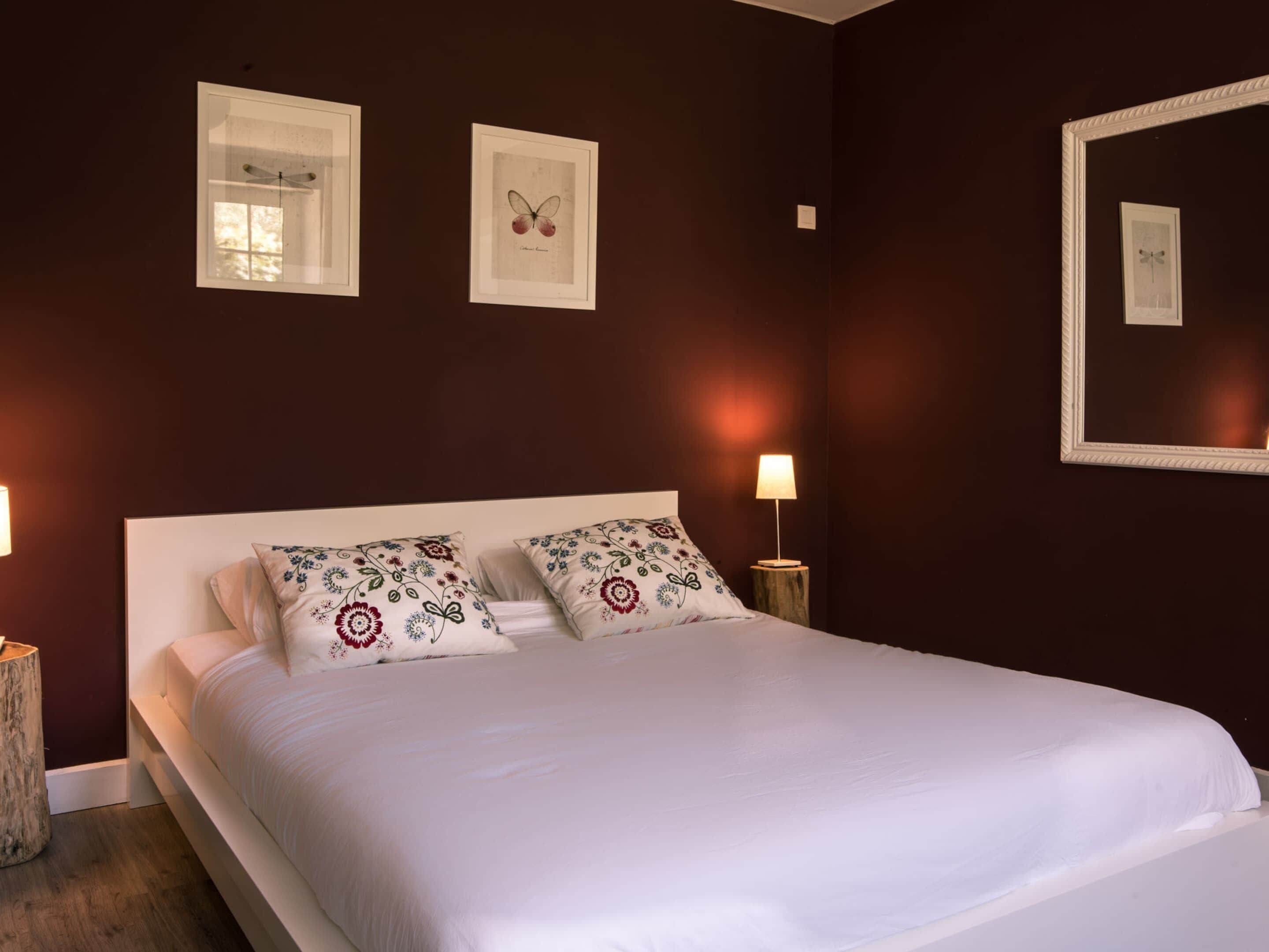 Una cama bien hecha con almohadas florales, una lámpara de noche y cuadros en la pared.