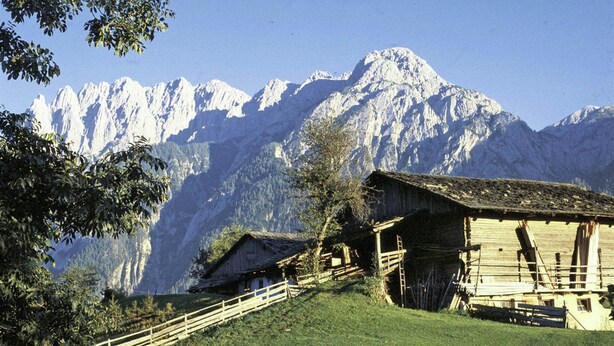 Uma cabana de montanha com fachada de madeira, cercada por uma cerca de madeira, e vista desobstruída de montanhas cobertas de neve ao fundo.