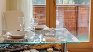 Mesa de jantar com tampo de vidro, posta com pratos, copos e talheres, com vista para um deck de madeira e vegetação.