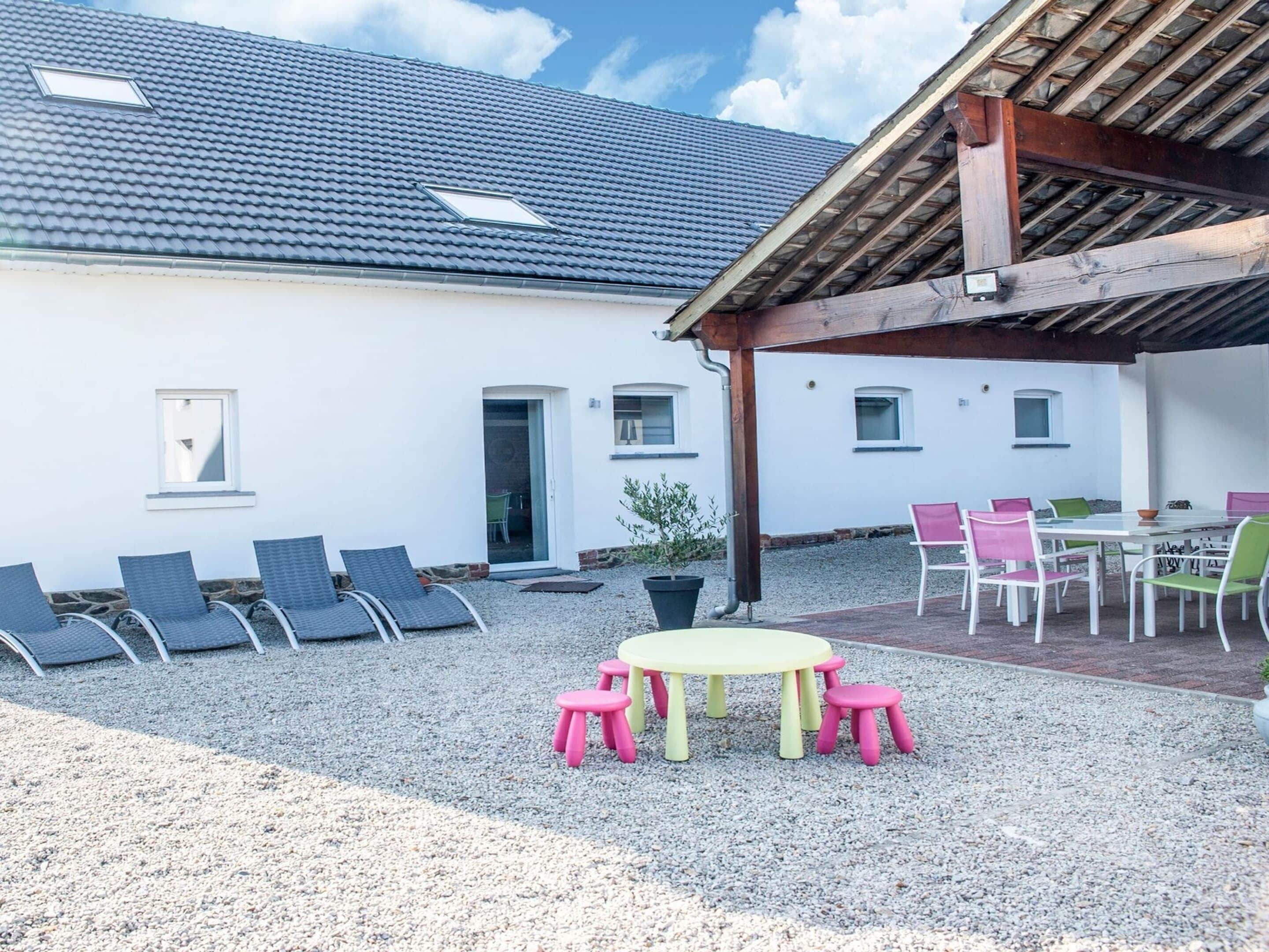 A covered outdoor seating area with a table and chairs, a gravel ground, and a building with a tiled roof and windows.