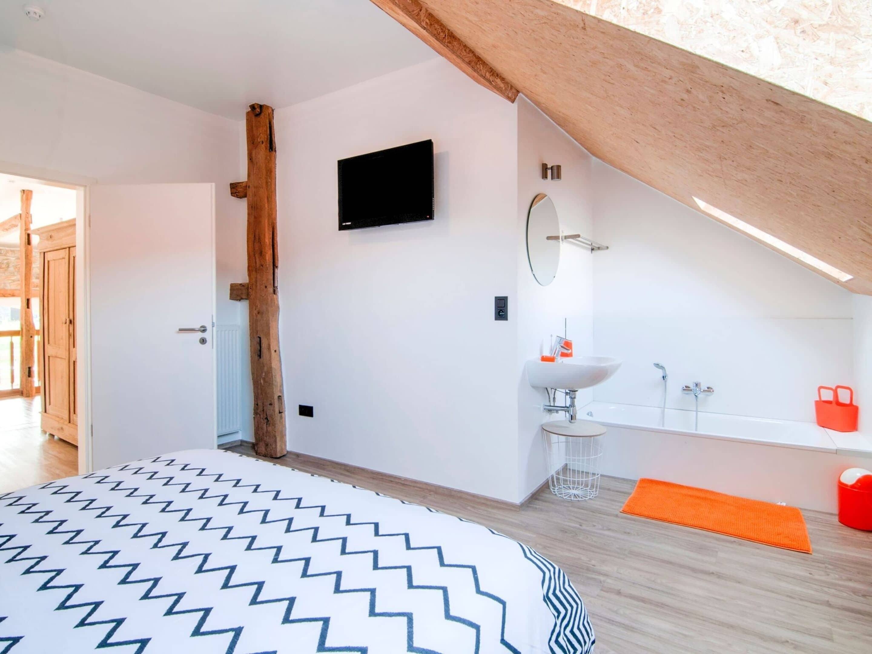 Un dormitorio con cama, televisor montado en la pared, baño con lavabo y bañera, y techo de madera.