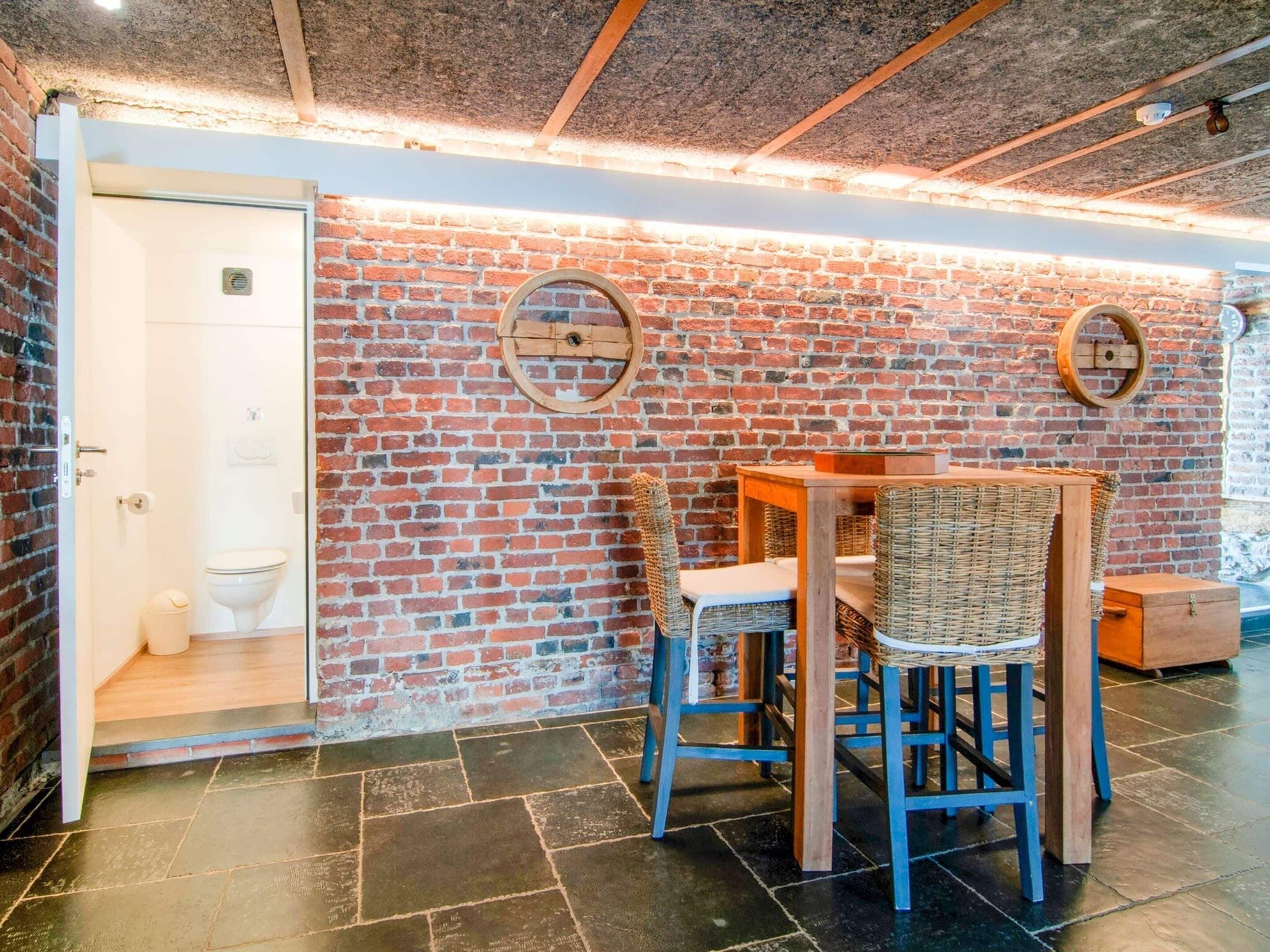 A bathroom with a brick wall, a toilet, and a sink. There is a table with chairs and a wooden cabinet.