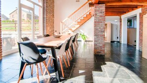 A spacious dining area with a long wooden table and modern chairs, a staircase with wooden railings, and a view of the outside through large windows. - Liege Villa Near Spa Circuit (Malmedy)
