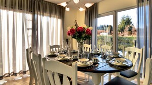 A dining room with a round table set for six, white chairs, a vase of flowers, and a view of the outdoors through large windows. - Wheelchair-friendly House With Sauna (Buetgenbach)