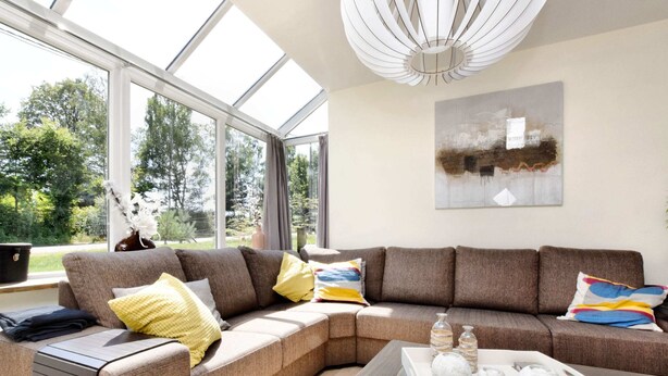 A modern living room with a sectional sofa, a glass coffee table, and a large window offering a view of greenery. - Wheelchair-friendly House With Sauna (Buetgenbach)