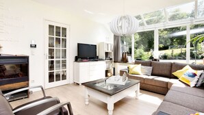 A modern living room with a sofa, coffee table, and TV. There is a bookshelf, a fireplace, and a large window with a view of greenery. - Wheelchair-friendly House With Sauna (Buetgenbach)