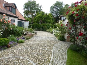 A cobblestone pathway leading through a garden with various flowering plants and a brick house with a tiled roof.