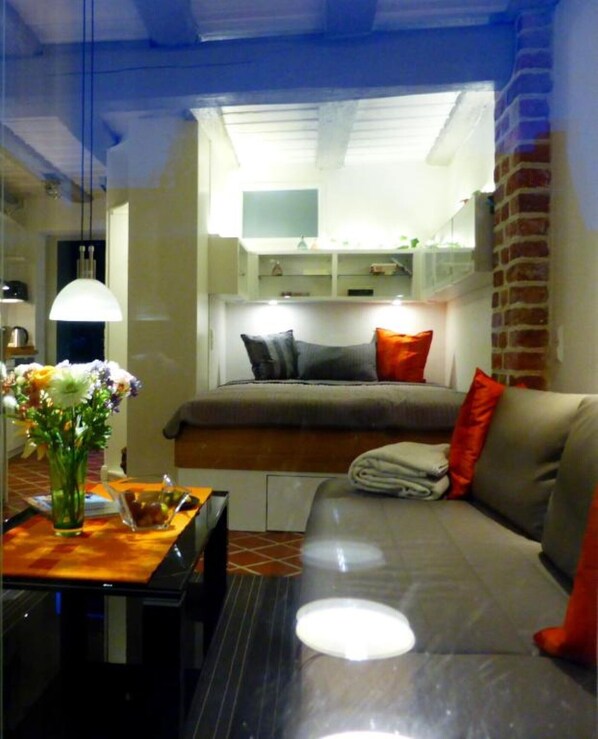 A compact room with a bed, a sofa, a table with a vase of flowers, and a hanging light fixture.
