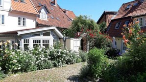 A cobblestone pathway leading through a garden with blooming flowers and a white building with a sloped roof. - Apartments gatehouse - old town (Lüneburg)