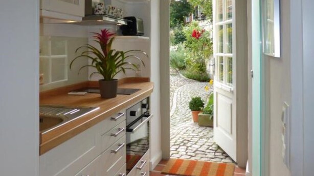 A narrow kitchen with white cabinets, a wooden countertop, and a potted plant. A door leads to a garden with a red flower and a tiled path. - Apartments gatehouse - old town (Lüneburg)