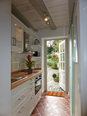 A narrow kitchen with white cabinets, a wooden countertop, and a potted plant. A door leads to a garden with a red flower and a tiled path.