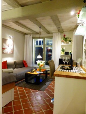 A living room with a sofa, coffee table, and a dining area with a table set for two. There is a window with curtains, a plant on a shelf, and a tiled floor.