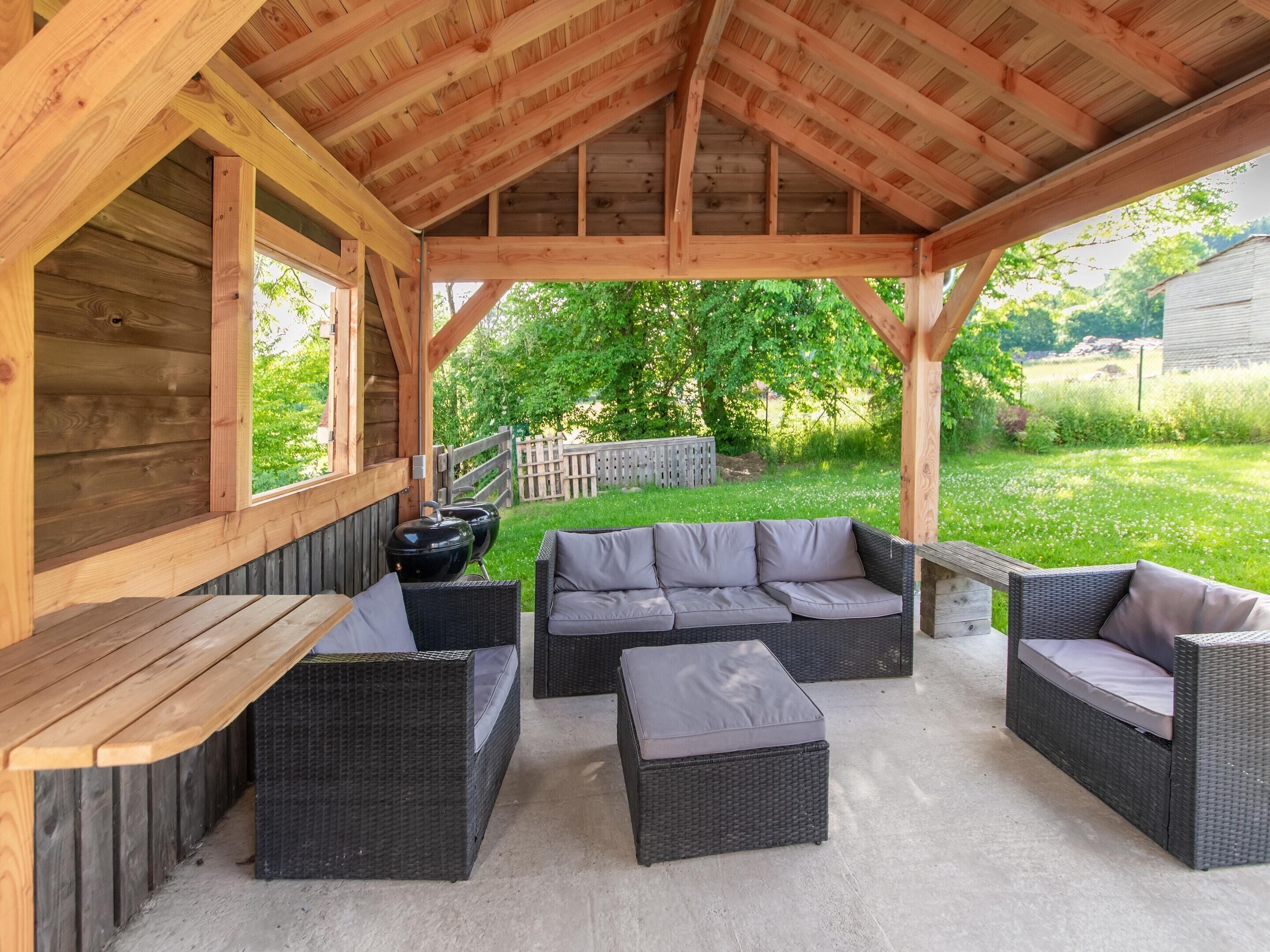 Une terrasse extérieure couverte, avec des poutres en bois, des meubles en osier et un barbecue.