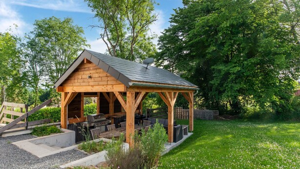 A wooden gazebo with a grill and seating area, surrounded by a garden with potted plants and a grassy lawn. - Villa in Freyneux With Garden (Manhay)