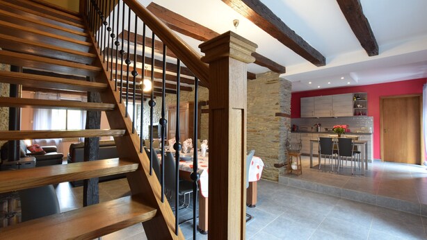 Un escalier en bois avec une rampe en métal mène à un espace repas agrémenté d’un mur en pierre et où l’on aperçoit une cuisine au fond.