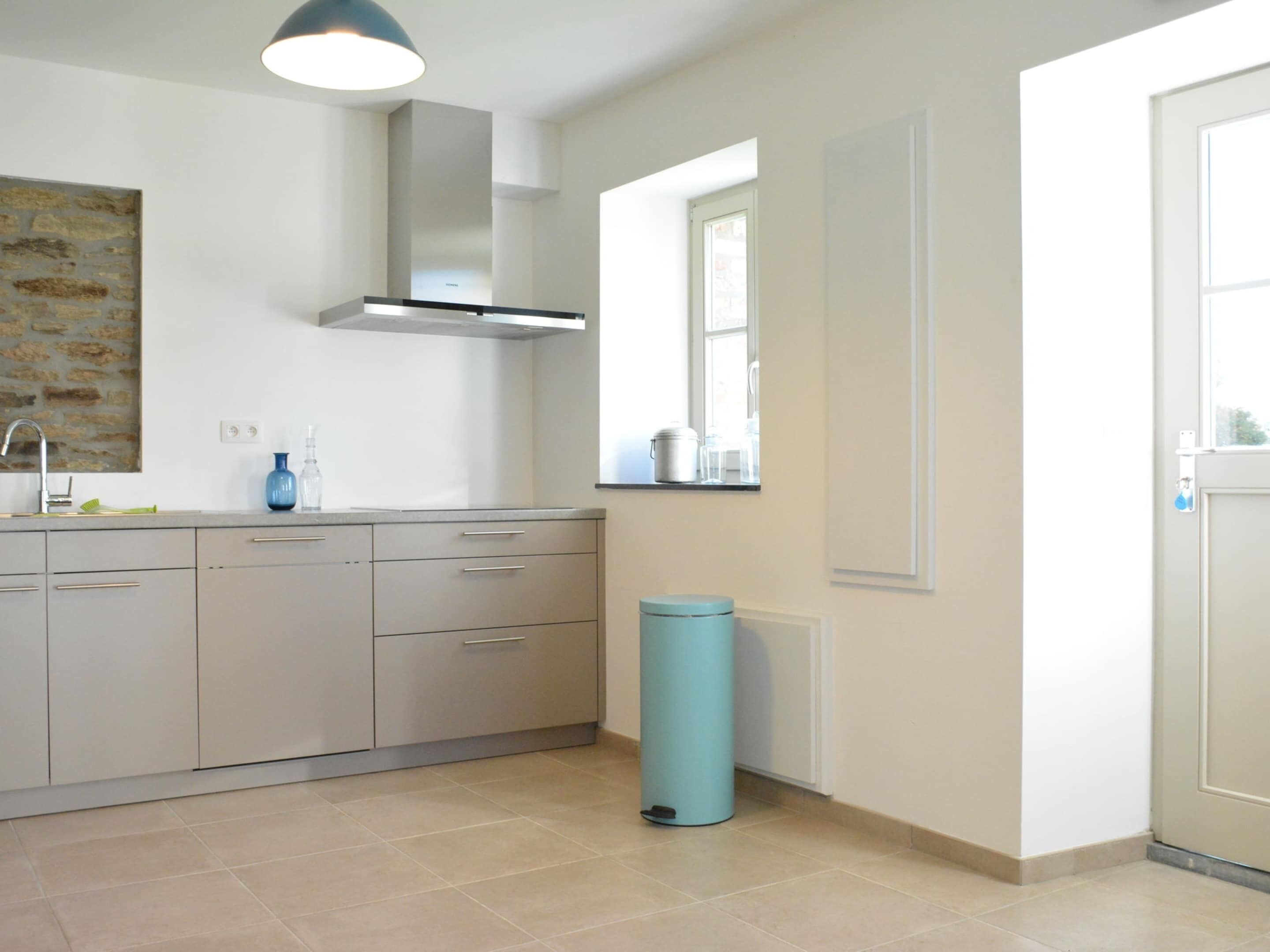 A modern kitchen with grey cabinets, a stainless steel range hood, a blue trash bin, and a window with a view of the outside.
