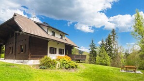 A two-story house with a sloped roof, a wooden deck, and a bench in the front yard. - Very Spacious, Detached Holiday Home in Carinthia near Skiing & Lakes (Gnesau)