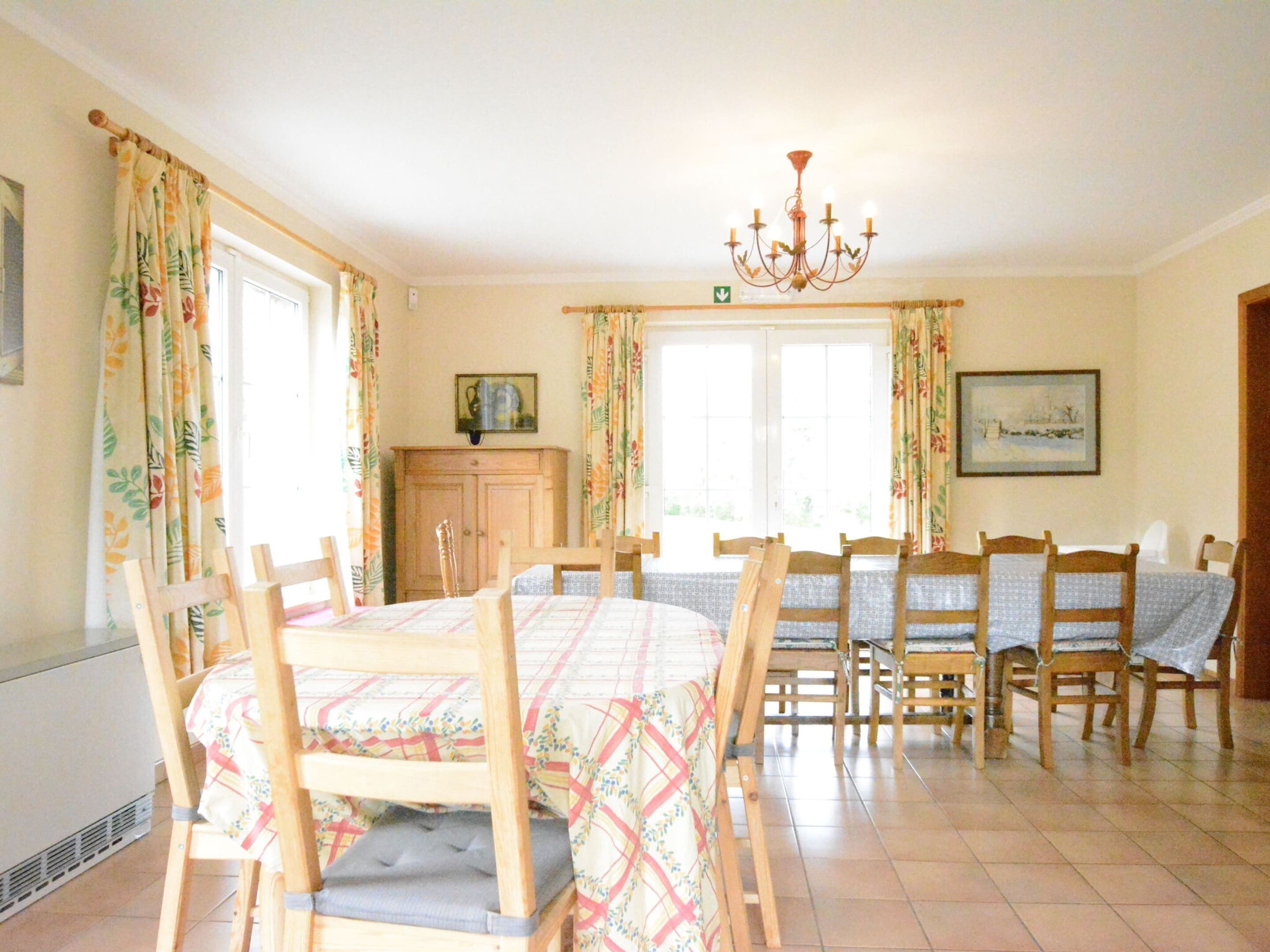 Uma sala de jantar com uma mesa posta para quatro pessoas, cadeiras de madeira, um candelabro e uma janela com cortinas.