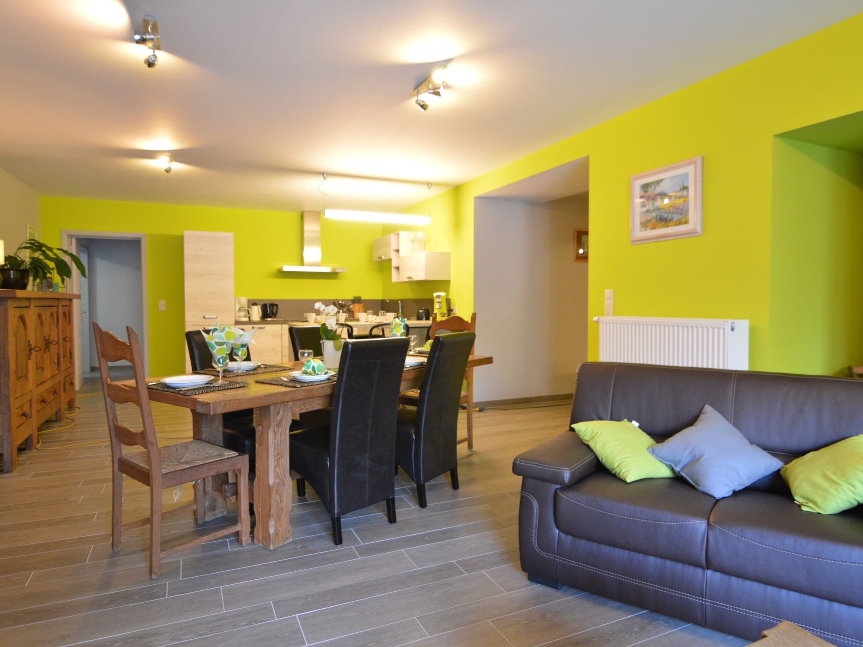 A modern living room with a brown leather sofa, a dining table set for four, and a kitchen in the background.