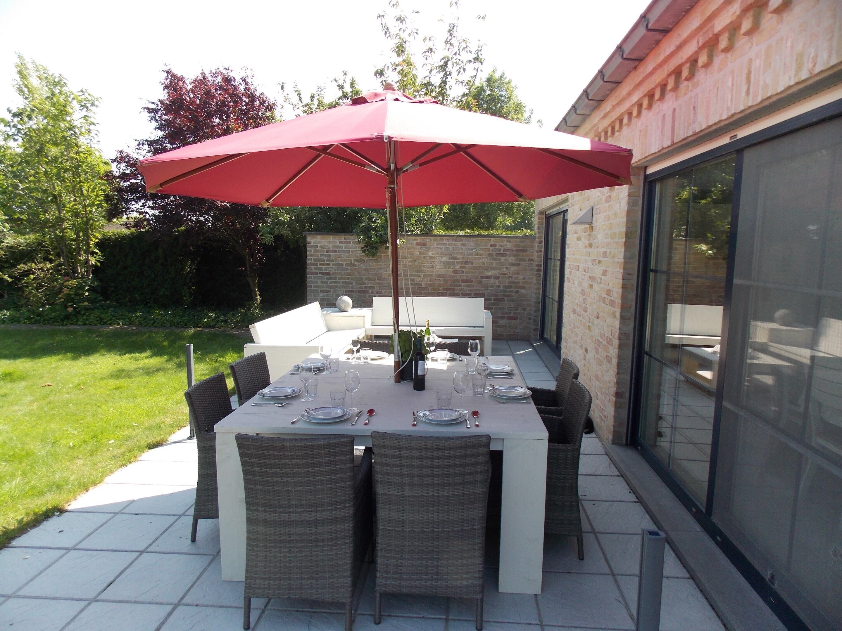 Une terrasse extérieure avec une table dressée pour le repas, entourée de chaises en osier, sous un parasol rouge.