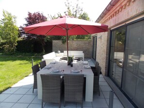 An outdoor patio with a table set for dining, surrounded by wicker chairs, under a red umbrella.