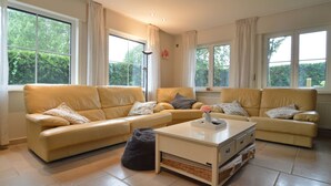 Een ruime woonkamer met twee beige leren banken, een witte salontafel en een groot raam met uitzicht op groen buiten.