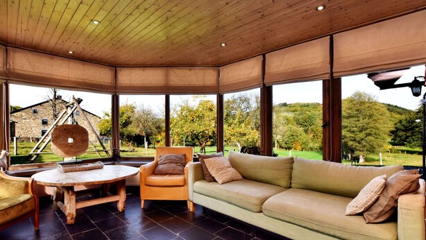 A cozy room with a wooden ceiling, a green sofa, a wooden table, and a chair. Large windows offer a view of a garden and a stone building. - Holiday Home in Vielsalm (Vielsalm)