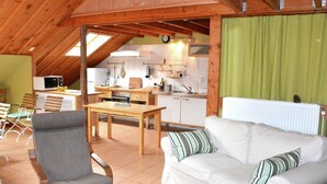 A cozy, open-plan living and kitchen area with wooden beams, a dining table, a sofa, and a kitchen with basic appliances.
