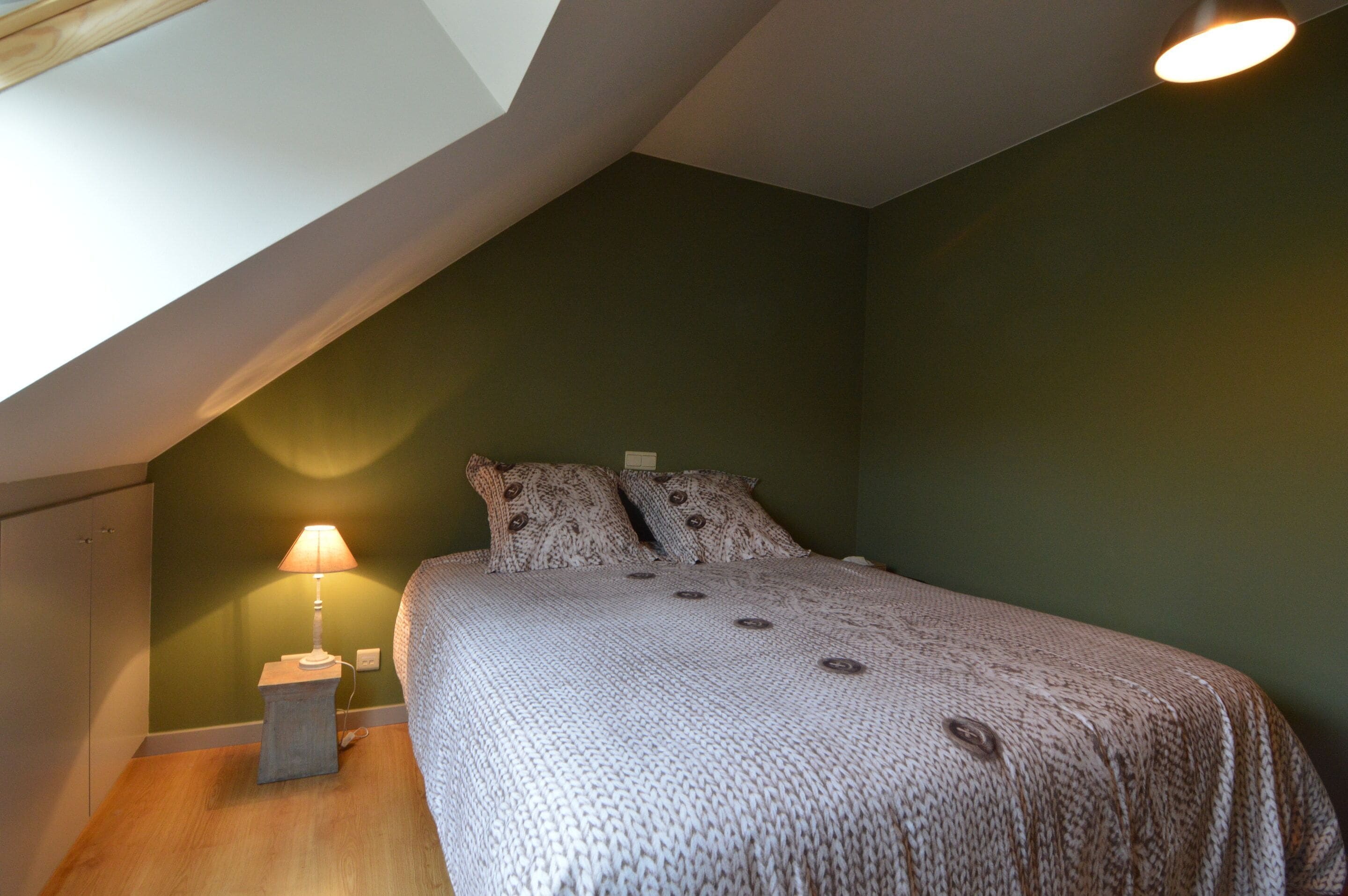 A bedroom with a sloped ceiling, a bed with patterned bedding, a bedside lamp, and a wall socket.
