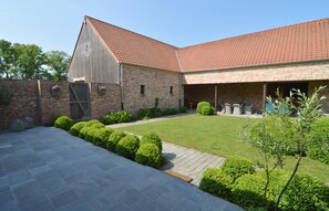 Un bâtiment en briques surmonté d’un toit de tuiles rouges, doté d’une terrasse pavée et de buissons soigneusement taillés.