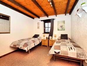 Une chambre avec deux lits simples, des têtes de lit en bois, une fenêtre avec des rideaux, une petite table avec une lampe et une étagère fixée au mur.