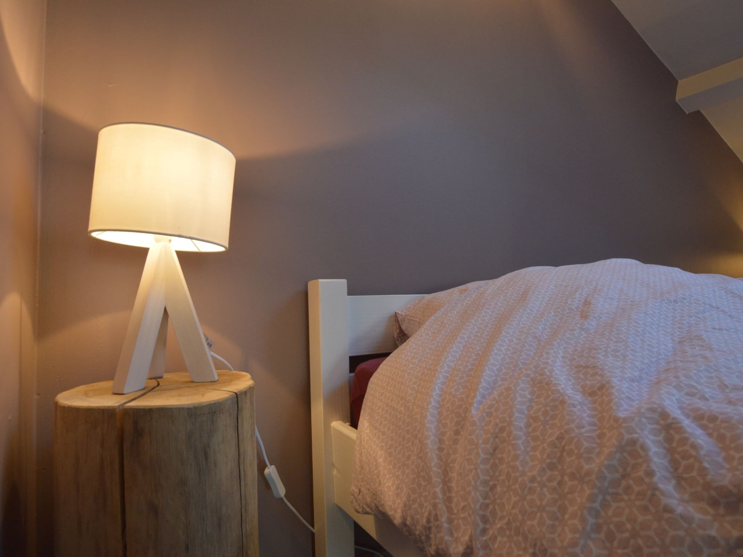 Un lit recouvert d’une couverture à motifs, une lampe de chevet posée sur une table de nuit en bois et une prise électrique sur le mur.